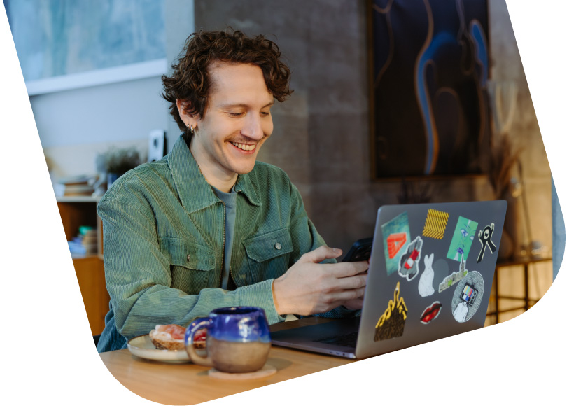 A young and smiling man is sitting at a table with an open computer and looking at his phone.