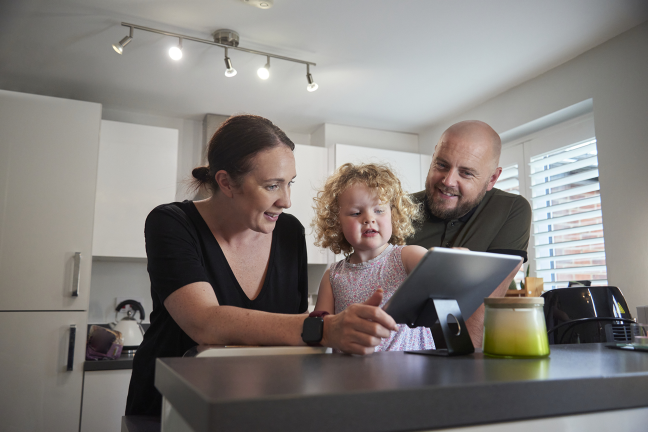 Smiling, a father and mother watch their young daughter use a tablet to control their smart home.